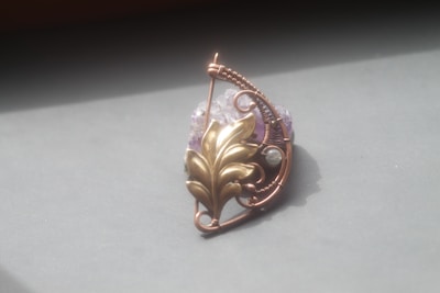 An intricately designed metal pendant with a swirling pattern and leaf motif, set against a muted background. The pendant incorporates a blend of copper wire and a brass-colored leaf shape, along with a textured purple stone.
