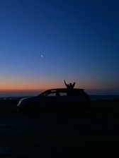 A silhouette of a car with a person raising their arm through the sunroof set against the backdrop of a twilight sky. The crescent moon and a star are visible in the deep blue sky, transitioning into shades of orange near the horizon.