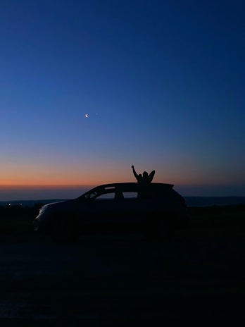 A silhouette of a car with a person raising their arm through the sunroof set against the backdrop of a twilight sky. The crescent moon and a star are visible in the deep blue sky, transitioning into shades of orange near the horizon.