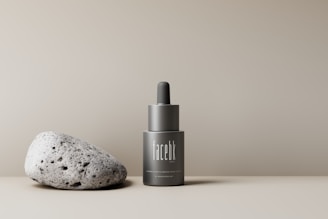 A minimalist setup featuring a gray, sleek bottle with a dropper cap labeled 'facebk' next to a textured, porous stone. The background is a neutral beige, creating a calming and sophisticated aesthetic.