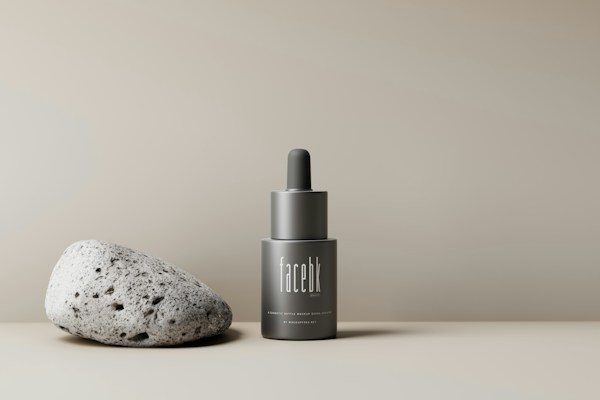 A minimalist setup featuring a gray, sleek bottle with a dropper cap labeled 'facebk' next to a textured, porous stone. The background is a neutral beige, creating a calming and sophisticated aesthetic.