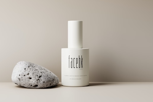 A minimalist scene featuring a white cylindrical bottle with the text 'facebk'. The bottle is positioned next to a textured, light-colored stone, both set against a soft beige background.