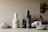 A collection of skincare bottles with minimalist designs arranged alongside textured stones against a muted background. The bottles vary in size and are labeled with 'facebk.' The scene is complemented by the presence of a small leafy plant, adding a touch of greenery.