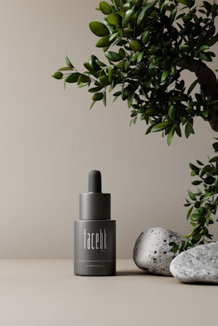 A minimalistic scene featuring a gray cosmetic bottle with a dropper top, labeled 'facebk'. It is set against a neutral beige background. A lush green plant is visible on the right side, accompanied by a couple of textured gray stones at the base, creating an elegant and natural aesthetic.