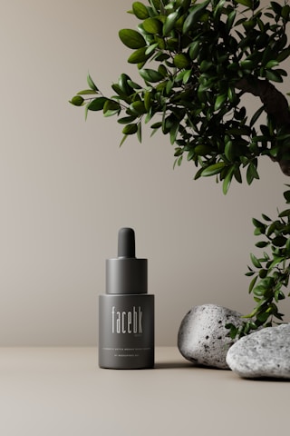 A minimalistic scene featuring a gray cosmetic bottle with a dropper top, labeled 'facebk'. It is set against a neutral beige background. A lush green plant is visible on the right side, accompanied by a couple of textured gray stones at the base, creating an elegant and natural aesthetic.