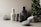 A minimalist arrangement of skincare product bottles in neutral tones is set against a soft beige background. The bottles are labeled 'facebk' and vary in height and size. Natural stone elements and lush green foliage accentuate the scene, adding a contrast of texture and color.
