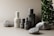 A minimalist arrangement of skincare product bottles in neutral tones is set against a soft beige background. The bottles are labeled 'facebk' and vary in height and size. Natural stone elements and lush green foliage accentuate the scene, adding a contrast of texture and color.