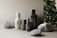 A minimalist arrangement of skincare product bottles in neutral tones is set against a soft beige background. The bottles are labeled 'facebk' and vary in height and size. Natural stone elements and lush green foliage accentuate the scene, adding a contrast of texture and color.