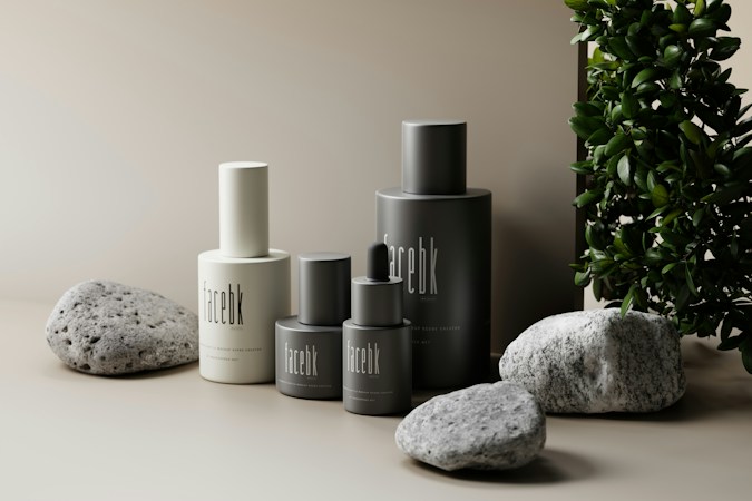 A minimalist arrangement of skincare product bottles in neutral tones is set against a soft beige background. The bottles are labeled 'facebk' and vary in height and size. Natural stone elements and lush green foliage accentuate the scene, adding a contrast of texture and color.