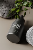 A dark green cylindrical cosmetic bottle displayed on its side, with the label 'facebk' in white. It is surrounded by lush green leaves and textured gray stones, set against a neutral background.