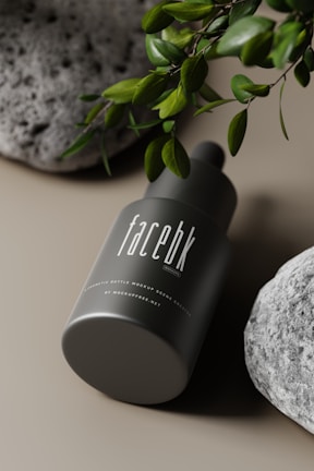 A dark green cylindrical cosmetic bottle displayed on its side, with the label 'facebk' in white. It is surrounded by lush green leaves and textured gray stones, set against a neutral background.