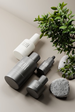 A collection of minimalist skincare or cosmetic bottles in sleek black and white designs. These are arranged on a soft, neutral-colored surface with a green plant branch and a few decorative stones placed nearby, adding a natural element to the composition.