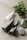 A collection of minimalist skincare or cosmetic bottles in sleek black and white designs. These are arranged on a soft, neutral-colored surface with a green plant branch and a few decorative stones placed nearby, adding a natural element to the composition.