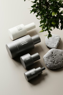 A minimalist arrangement featuring several cylindrical cosmetic bottles in various sizes, each labeled with a modern, sleek font, alongside two textured stones and some lush green leaves over a neutral, light-colored background.