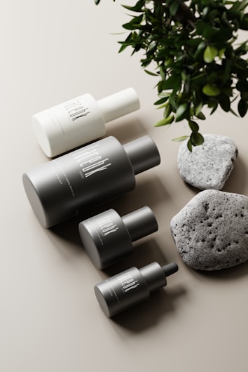A minimalist arrangement featuring several cylindrical cosmetic bottles in various sizes, each labeled with a modern, sleek font, alongside two textured stones and some lush green leaves over a neutral, light-colored background.