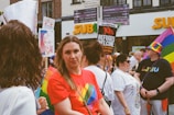 A group of people gathered in an outdoor setting, many wearing colorful clothing and accessories, including rainbow patterns. One person holds a sign that reads 'Refugees Welcome.' Another individual is wearing a colorful rainbow hat.