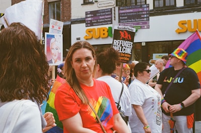 A group of people gathered in an outdoor setting, many wearing colorful clothing and accessories, including rainbow patterns. One person holds a sign that reads 'Refugees Welcome.' Another individual is wearing a colorful rainbow hat.