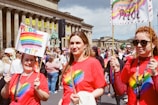 A group of people are participating in a pride parade, wearing red t-shirts featuring a rainbow heart design. They are holding signs with rainbow colors, and there is a festive crowd around them. The backdrop includes a historic building with columns and banners.