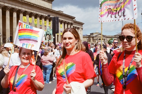 A group of people are participating in a pride parade, wearing red t-shirts featuring a rainbow heart design. They are holding signs with rainbow colors, and there is a festive crowd around them. The backdrop includes a historic building with columns and banners.