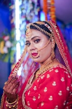 A woman dressed in traditional bridal attire with intricate jewelry, including a nose ring and headpiece. She wears a red embroidered outfit, and her hands are adorned with mehndi designs. The background is blurred with colorful decorations.