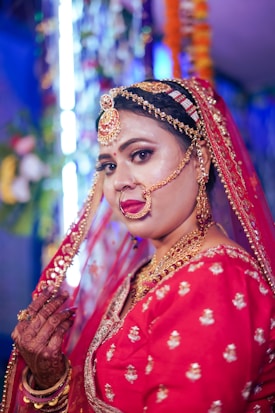A woman dressed in traditional bridal attire with intricate jewelry, including a nose ring and headpiece. She wears a red embroidered outfit, and her hands are adorned with mehndi designs. The background is blurred with colorful decorations.