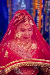 A person wearing a traditional red wedding attire with intricate gold embroidery and jewelry. The individual is adorned with a bright red veil, ornate headpiece, and lavish necklaces. The background features colorful and blurred bokeh lights.