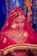 A person wearing a traditional red wedding attire with intricate gold embroidery and jewelry. The individual is adorned with a bright red veil, ornate headpiece, and lavish necklaces. The background features colorful and blurred bokeh lights.