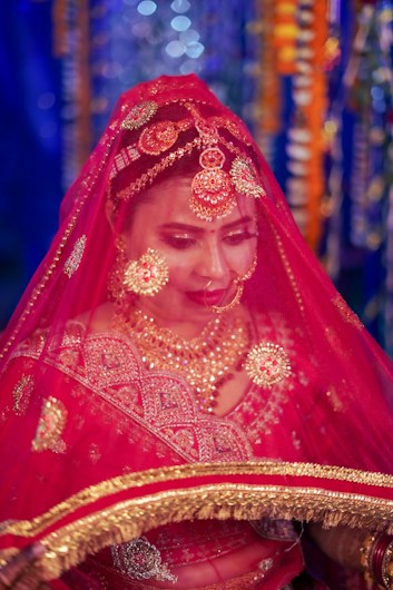 A person wearing a traditional red wedding attire with intricate gold embroidery and jewelry. The individual is adorned with a bright red veil, ornate headpiece, and lavish necklaces. The background features colorful and blurred bokeh lights.