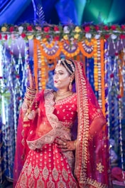 A woman is dressed in a traditional, ornate red bridal outfit with intricate embroidery and gold jewelry. She stands in front of a decorated backdrop featuring colorful garlands and flowers, creating a festive and vibrant setting. The lighting highlights her elaborate attire and accessories.