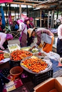 A vibrant market scene with vendors and buyers engaged in activities. Stalls are filled with fresh produce, particularly oranges, displayed in baskets. People are dressed in colorful traditional attire, with some wearing hats. The setting is under a rustic, covered area with wooden beams and corrugated metal roofing.