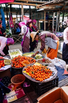 A vibrant market scene with vendors and buyers engaged in activities. Stalls are filled with fresh produce, particularly oranges, displayed in baskets. People are dressed in colorful traditional attire, with some wearing hats. The setting is under a rustic, covered area with wooden beams and corrugated metal roofing.