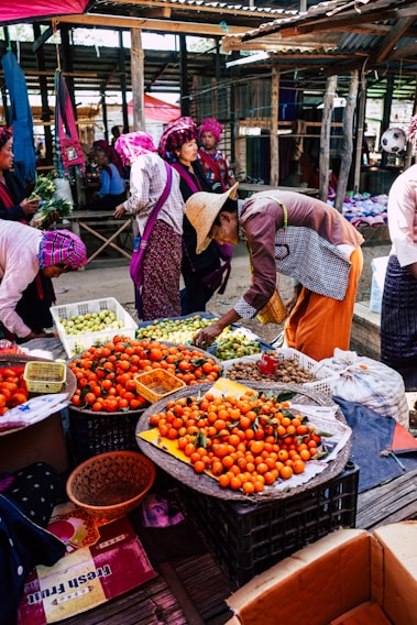 A vibrant market scene with vendors and buyers engaged in activities. Stalls are filled with fresh produce, particularly oranges, displayed in baskets. People are dressed in colorful traditional attire, with some wearing hats. The setting is under a rustic, covered area with wooden beams and corrugated metal roofing.