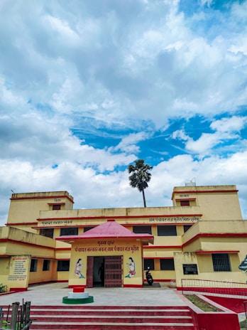 A two-story yellow and red building with a red entrance canopy and text in Hindi above it. The building is situated under a partly cloudy blue sky. A single tall palm tree can be seen behind the building.