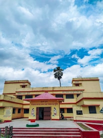 A two-story yellow and red building with a red entrance canopy and text in Hindi above it. The building is situated under a partly cloudy blue sky. A single tall palm tree can be seen behind the building.