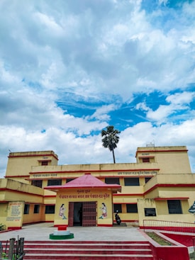 A two-story yellow and red building with a red entrance canopy and text in Hindi above it. The building is situated under a partly cloudy blue sky. A single tall palm tree can be seen behind the building.