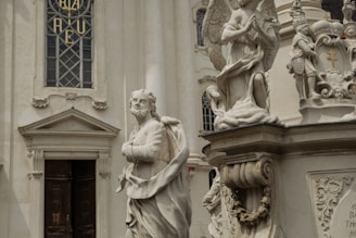 A detailed marble statue of a robed figure stands in front of an ornate building facade. The background includes architectural elements like a stained glass window with intricate designs, and another statue of an angel kneeling in prayer. The stone sculptures exhibit delicate craftsmanship and classical elements.