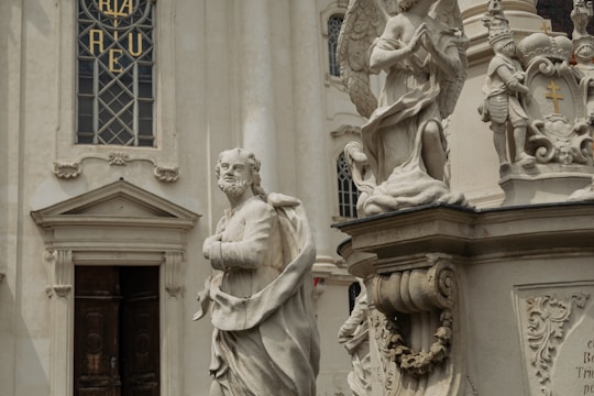 A detailed marble statue of a robed figure stands in front of an ornate building facade. The background includes architectural elements like a stained glass window with intricate designs, and another statue of an angel kneeling in prayer. The stone sculptures exhibit delicate craftsmanship and classical elements.
