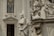 A detailed marble statue of a robed figure stands in front of an ornate building facade. The background includes architectural elements like a stained glass window with intricate designs, and another statue of an angel kneeling in prayer. The stone sculptures exhibit delicate craftsmanship and classical elements.
