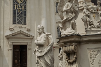 A detailed marble statue of a robed figure stands in front of an ornate building facade. The background includes architectural elements like a stained glass window with intricate designs, and another statue of an angel kneeling in prayer. The stone sculptures exhibit delicate craftsmanship and classical elements.