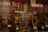 A display window showcasing a collection of religious figurines, including depictions of saints, Mary holding Jesus, and angels. The background reflects an urban street scene with pedestrian silhouettes faintly visible.