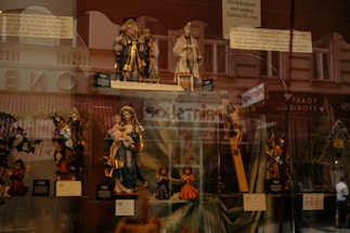 A display window showcasing a collection of religious figurines, including depictions of saints, Mary holding Jesus, and angels. The background reflects an urban street scene with pedestrian silhouettes faintly visible.