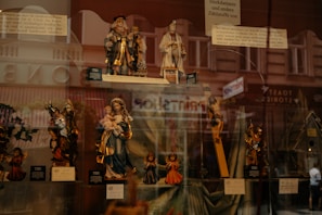 A display window showcasing a collection of religious figurines, including depictions of saints, Mary holding Jesus, and angels. The background reflects an urban street scene with pedestrian silhouettes faintly visible.