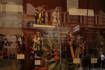 A display window showcasing a collection of religious figurines, including depictions of saints, Mary holding Jesus, and angels. The background reflects an urban street scene with pedestrian silhouettes faintly visible.