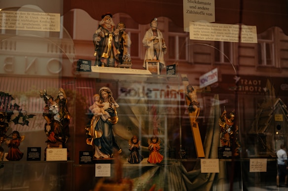 A display window showcasing a collection of religious figurines, including depictions of saints, Mary holding Jesus, and angels. The background reflects an urban street scene with pedestrian silhouettes faintly visible.