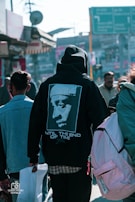 A person wearing a black hoodie with an image of a famous musician and the words 'Until The End of Time' on the back is walking in a busy outdoor urban setting. The street is bustling with multiple people, and there are shops and colorful signs in the background.