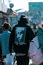 A person wearing a black hoodie with an image of a famous musician and the words 'Until The End of Time' on the back is walking in a busy outdoor urban setting. The street is bustling with multiple people, and there are shops and colorful signs in the background.