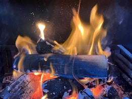 Logs of wood are burning with bright yellow and orange flames in a fireplace. The fire casts a warm glow over the dark charred wood, and small embers are visible at the base.