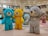 Large colorful teddy bear sculptures are displayed in a shopping mall. There are bears in various colors including blue, yellow, gray, mint, and pink, each standing on a patterned floor in an open area beneath a circular ceiling design. Shoppers and stores can be seen in the background.