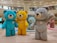 Large colorful teddy bear sculptures are displayed in a shopping mall. There are bears in various colors including blue, yellow, gray, mint, and pink, each standing on a patterned floor in an open area beneath a circular ceiling design. Shoppers and stores can be seen in the background.