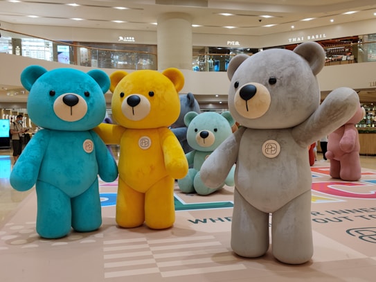 Large colorful teddy bear sculptures are displayed in a shopping mall. There are bears in various colors including blue, yellow, gray, mint, and pink, each standing on a patterned floor in an open area beneath a circular ceiling design. Shoppers and stores can be seen in the background.
